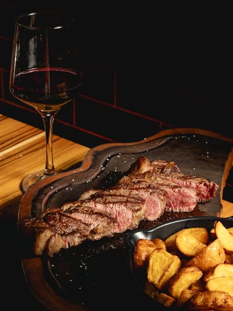 Corte de carne fileteado sobre una tabla, con copa de vino y papas